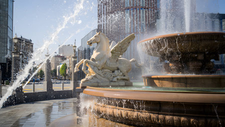 A stone Pegasus sculpture with wings is featured in a city square fountain with water splashing.の写真素材