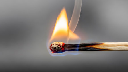 showing burning matchstick with bright orange flame and white smoke against a gray...