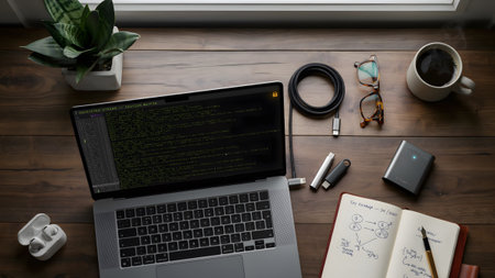 A top-down view of a wooden desk featuring a laptop with code, a notebook, and various tech tools.