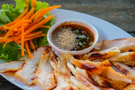 Grilled pork in a white dish with vegetables and dipping sauce on a wooden table.の写真素材