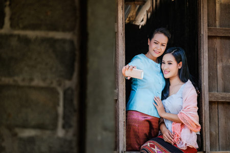 Two Mother and daughter in a traditional Thai dress are taking pictures of themselves with a mobile phone from a wooden window.の写真素材