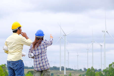An engineer stands holding a blueprint and looks at a wind turbine project to generate electricity and check wind direction.の写真素材