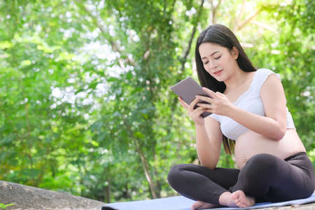 Asian 8 month pregnant woman using digital tablet sitting on grass in outdoor park. Outdoor nature background.の写真素材