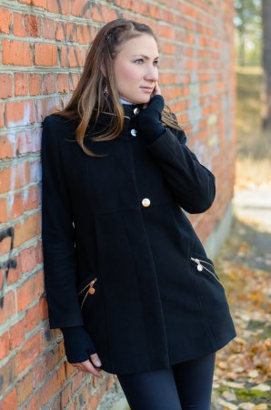 The beautiful girl in a black coat stands near a brick wall, having brought a palm to the face.の写真素材