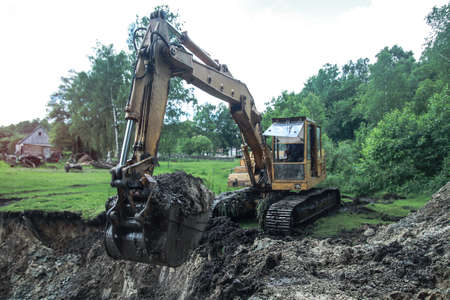 An excavator digs a large trench for building a house. A tractor digs a large lake that is already gaining water. Stock background for designの写真素材