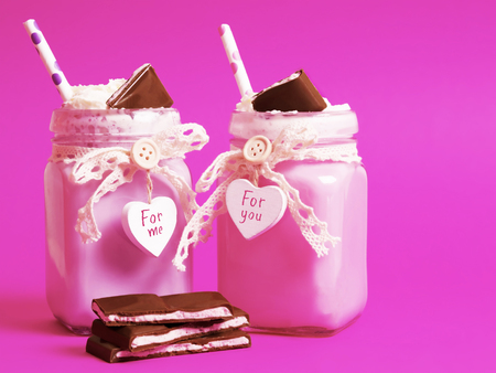 Delicious pink strawberry cocktail with ice cream with a whipped cream and chocolate in a beautiful jar with a heart on a pink background. There are slices of chocolate nearby.The inscriptions on the heartの写真素材