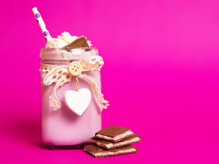 Delicious pink cocktail with ice cream with whipped cream and chocolate in a beautiful jar with a heart on a pink background. There are slices of chocolate nearby. Copy space.の写真素材