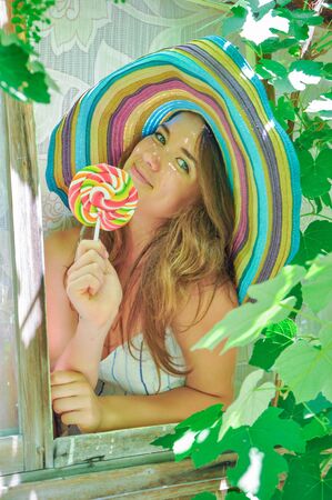 funny girl wearing a colorful hat with lollipop in a window with grape leaves in high qualityの写真素材