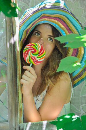 funny girl wearing a colorful hat with lollipop in a window with grape leaves in high qualityの写真素材