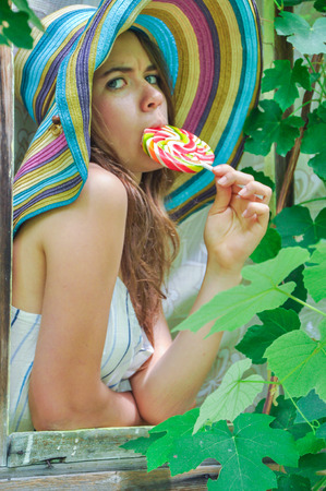 funny girl wearing a colorful hat with lollipop in a window with grape leaves in high qualityの写真素材
