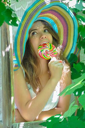 funny girl wearing a colorful hat with lollipop in a window with grape leaves in high qualityの写真素材