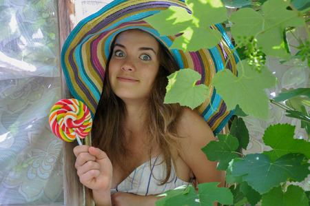 funny girl wearing a colorful hat with lollipop in a window with grape leaves in high qualityの写真素材
