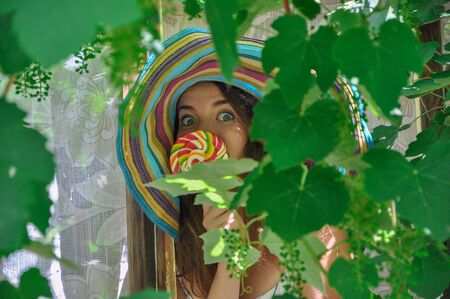 funny girl wearing a colorful hat with lollipop in a window with grape leaves in high qualityの写真素材