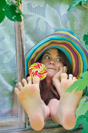 funny girl wearing a colorful hat with lollipop in a window with grape leaves in high qualityの写真素材