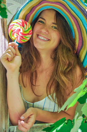 funny girl wearing a colorful hat with lollipop in a window with grape leaves in high qualityの写真素材