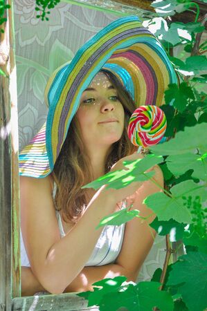 funny girl wearing a colorful hat with lollipop in a window with grape leaves in high qualityの写真素材