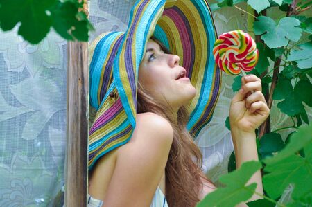 funny girl wearing a colorful hat with lollipop in a window with grape leaves in high qualityの写真素材