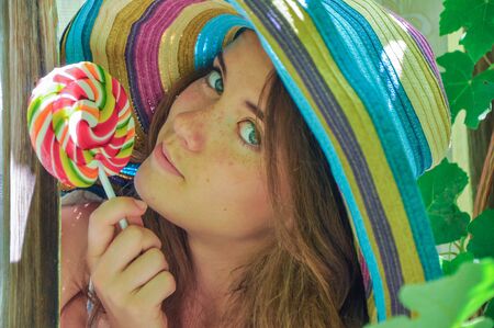 funny girl wearing a colorful hat with lollipop in a window with grape leaves in high qualityの写真素材