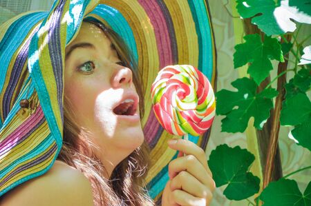 funny girl wearing a colorful hat with lollipop in a window with grape leaves in high qualityの写真素材