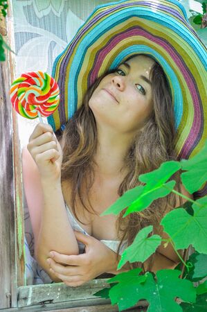 funny girl wearing a colorful hat with lollipop in a window with grape leaves in high qualityの写真素材