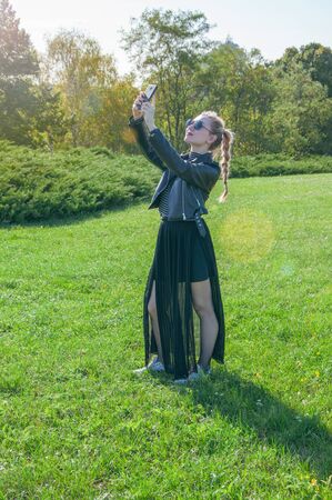 A beautiful blonde girl stands on a green lawn background and makes selfieの写真素材