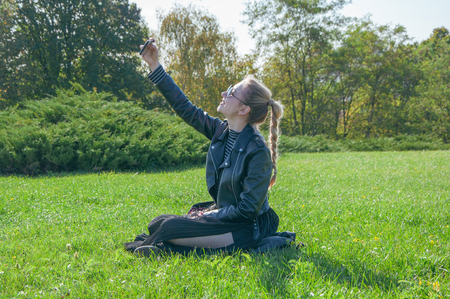 A beautiful blonde girl sitting on a green lawn and makes selfieの写真素材