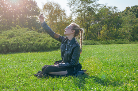 A beautiful blonde girl sitting on a green lawn and makes selfieの写真素材
