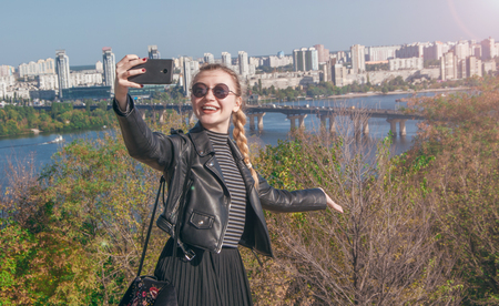 A beautiful blonde girl stands on the background of the city and makes selfieの写真素材