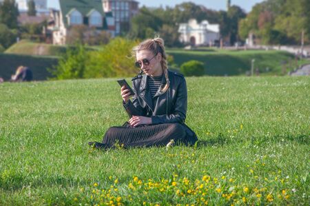 A beautiful blonde girl sits on a green lawn and looks in a smartphoneの写真素材