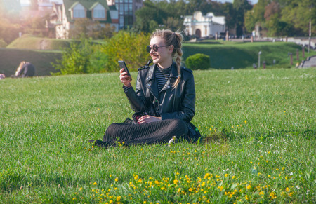 A beautiful blonde girl sits on a green lawn and looks in a smartphoneの写真素材