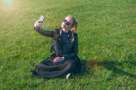 A beautiful blonde girl sitting on a green lawn and makes selfieの写真素材