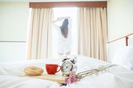 Closeup alarm clock having a good day with background happy woman stretching in bed after waking up, sunlight in morning.の写真素材