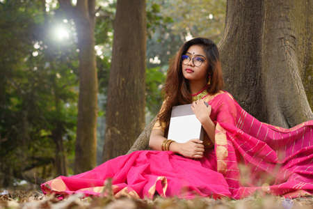 Outdoor portrait of pleased brunette beautiful Indian woman in eyeglasses wearing traditional Indian saree and jewellery, sitting under a tree and holding a book in park. Lifestyle and Fashion.の写真素材