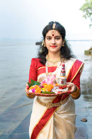 Portrait of beautiful Indian girl in front of ganga river wearing traditional Indian saree, gold jewellery and bangles holding plate of religious offering.の写真素材