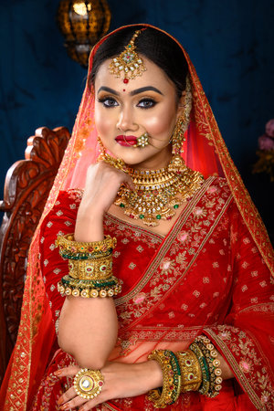 Stunning Indian bride dressed in traditional red bridal lehenga with heavy gold jewellery and veil sitting in a chair smiles tenderly in studio lighting. Wedding fashion and lifestyle.の写真素材