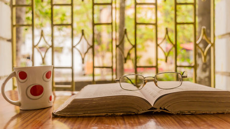 An open book a coffee cup and a specs or eyeglass on wooden table, warm sunny morning, side view...