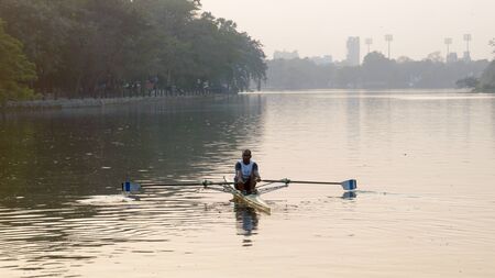 Individual Sports Speed Rower in Single scull crew rowing boat sliding racing shell on lake water oars in motion sitting sliding rigger seat doing exercise, fitness workout in sunset. Kolkata May 2019のeditorial素材