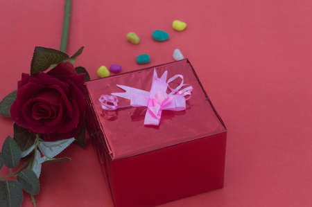 Close up gift box with tied bow ribbon placed near red rose on a table near in red background. High angle view. Design for valentine's day, anniversary or holiday celebration decoration.の写真素材