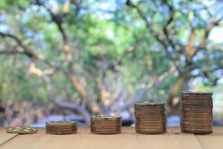Coin pile stacked in increasing chart shape on wood board with out of focus big tall green trees canopy background. Environmental friendly economy. Finance business of saving nature concept.の写真素材