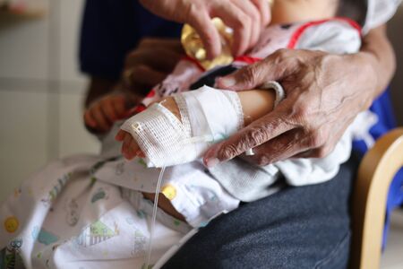 saline infusion. sick baby inward patient sitting on parent legs. baby in hospital being fed. sick kid in treatment process, in hospital and caring by many people.の写真素材