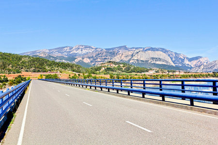 Modern Road Bridge Bordered by Blue Railings Crosses the Wide River Towards Farms and the Mountains Beyondの写真素材