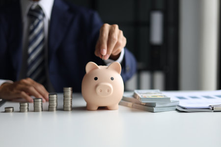 AÂ person in a suit putting a coin into a piggy bank with stacks of coins and money on the table. Saving money and financial planning, financial growth, investment, and future planning concept.の写真素材