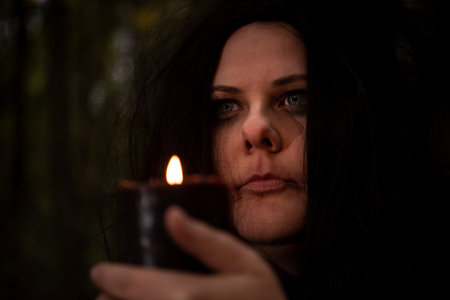 Black-haired witch holding candle in her hands in the forest.