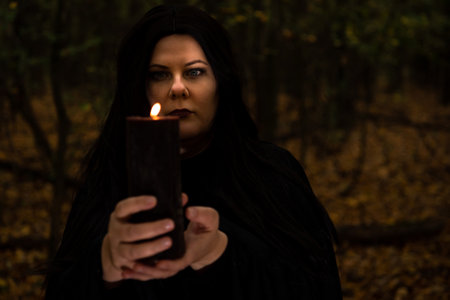 Black-haired witch holding candle in her hands in the forest.の写真素材