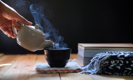 Pouring hot tea in teapot and cup on wood table, black background. Winter tea time.の写真素材