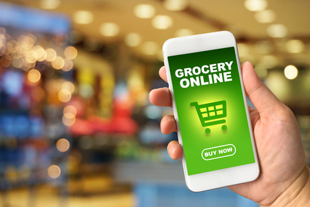 Woman hand holding smartphone against blur bokeh of store background Grocery online conceptの写真素材