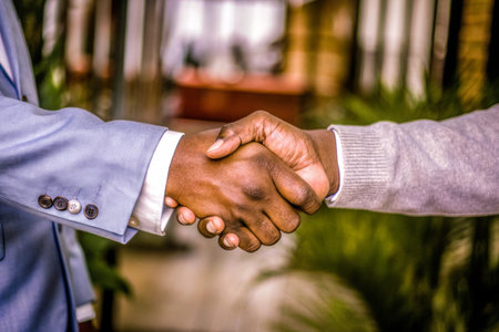 This eye-level shot captures a business handshake between two men wearing professional attire against a blurred background of greenery. It evokes themes of partnership, agreement, and the successful completion of a deal.の素材