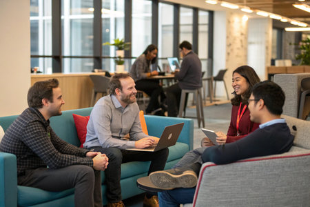 A diverse group of professionals sit in a modern office lounge, collaborating on laptops and tablets, sharing ideas and solutions. The scene showcases a modern and collaborative work environment, ideal for depicting teamwork, technology use, and workplace diversity.の素材