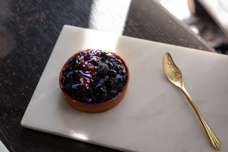 The blueberry pie on a white plate made of marble and a golden spoon has the sunlight shining on the...