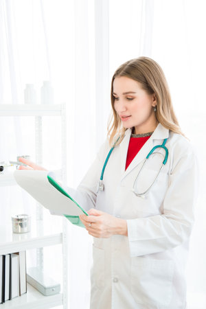 A female beautiful doctor is wearing a doctor uniform have a stethoscope and holding a green paper chart, smiling and looking at the camera the background is a white curtain in the hospital or clinic.の写真素材